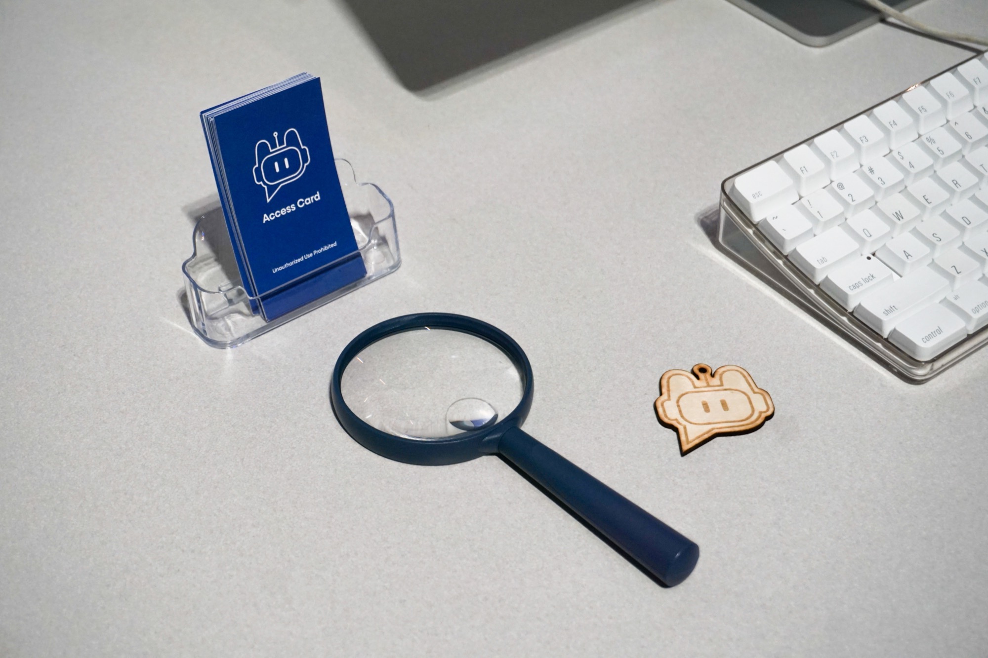 A blue magnifying glass, a wooden keychain shaped like a cartoon face, a stack of blue access cards in a holder, and a white computer keyboard on a light gray desk.