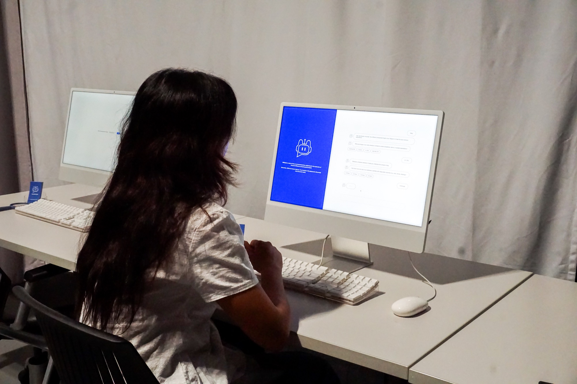 A person sits at a desk using a computer, viewing a screen with a chat interface and a blue section displaying a chatbot icon; another similar computer is nearby.