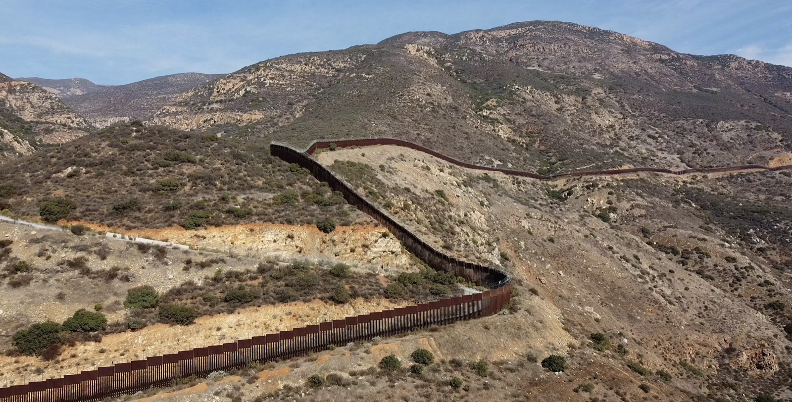 A tall metal border fence winds along rugged, mountainous terrain with brown hills and sparse vegetation under a blue sky.