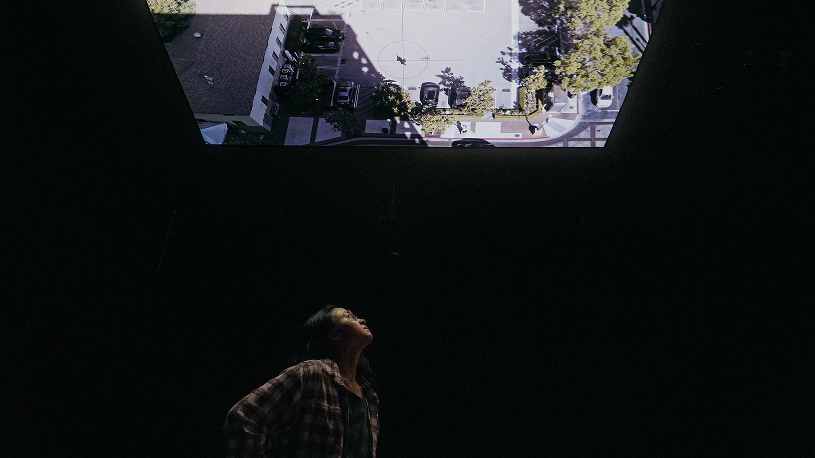 A person in a plaid shirt stands in darkness, looking up at a brightly lit, rectangular projection of an aerial view of a parking lot with cars and trees above them.