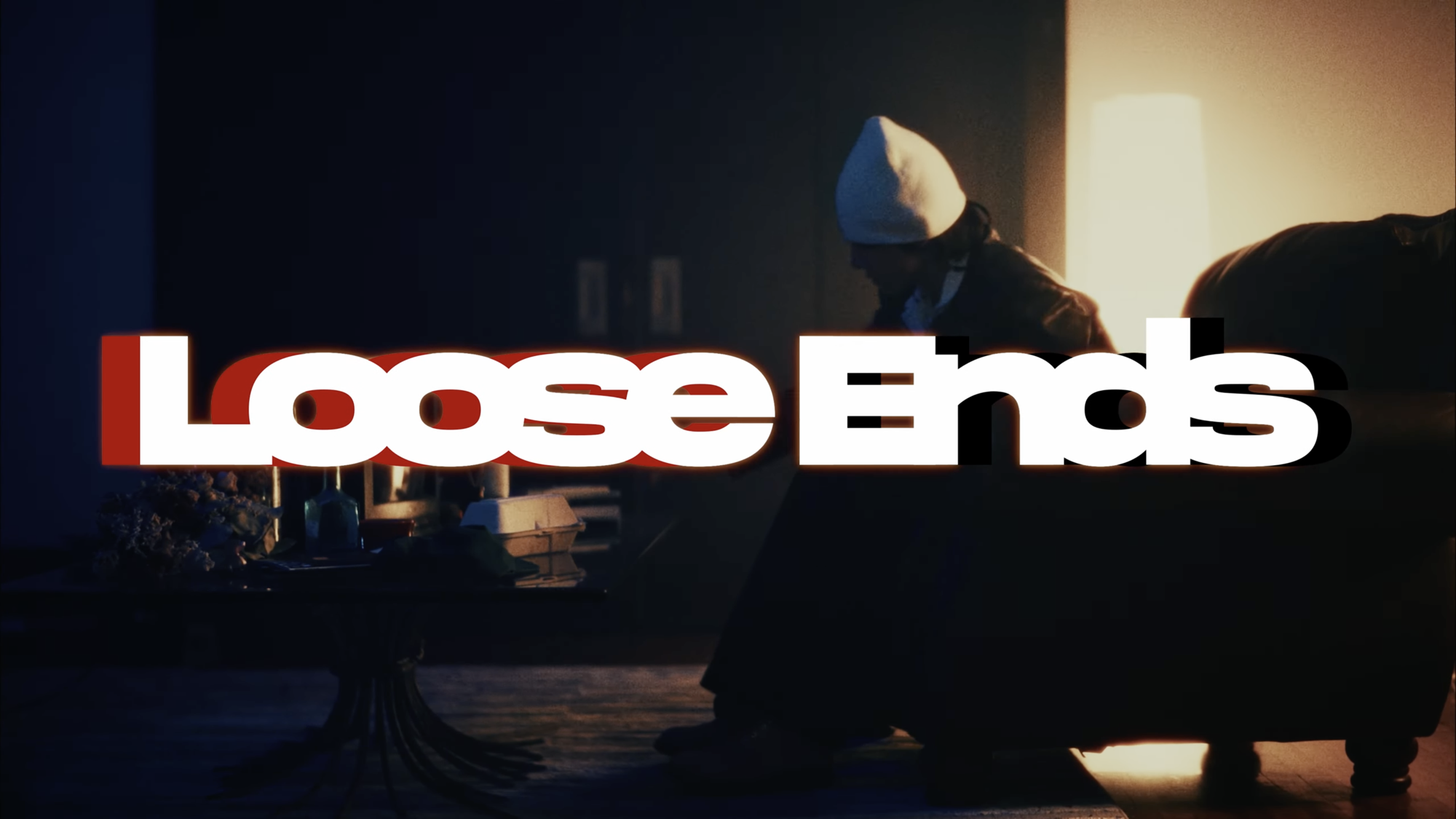 A person in a white beanie sits on a couch in a dimly lit room with a lamp and table nearby. Large text reading Loose Ends in bold white and red font is overlaid across the center.
