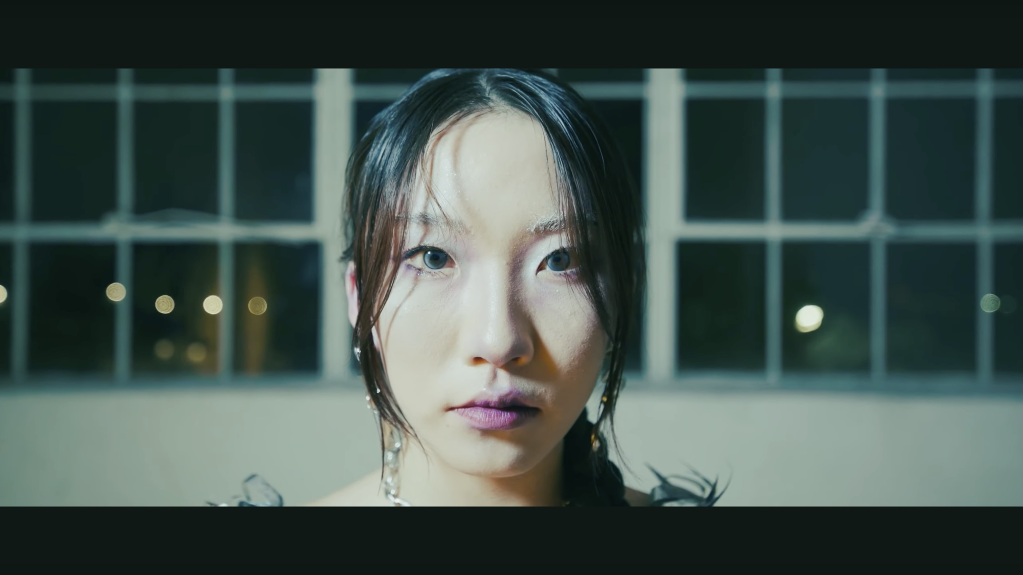 A person with long dark hair and light makeup looks directly at the camera in a well-lit indoor setting. Large windows are visible in the background with blurred lights outside.