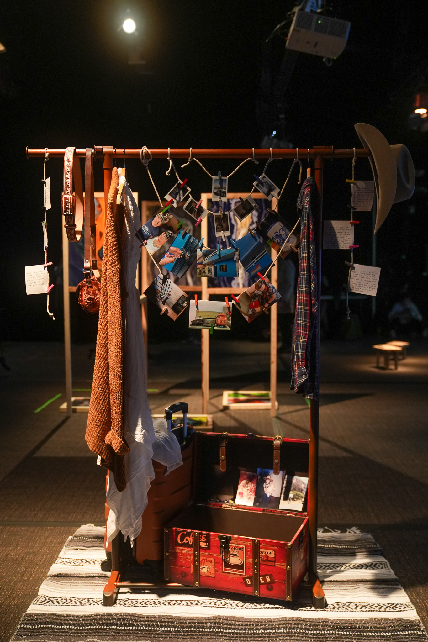 A wooden clothing rack displays hats, belts, clothes, and hanging photos and notes. At its base sits an open vintage suitcase filled with books and keepsakes, all set on a striped rug in a dimly lit room.