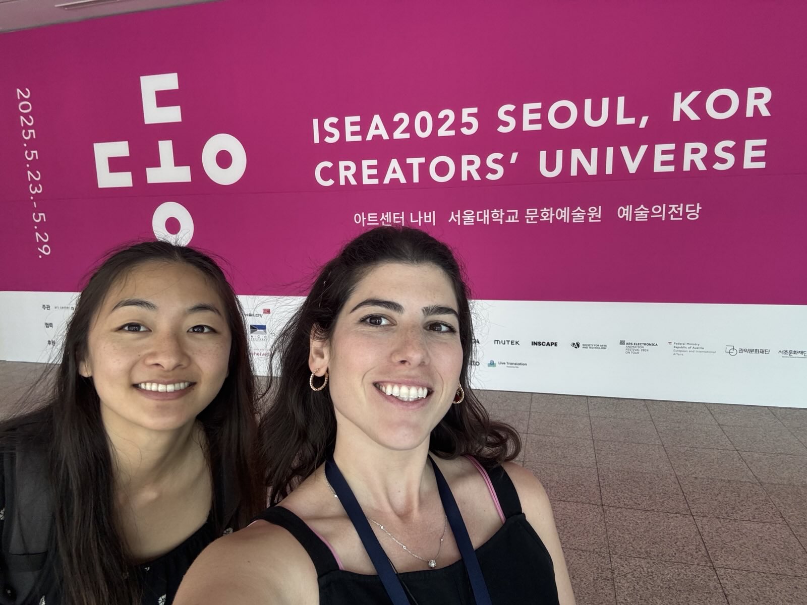 Two people smile for a selfie in front of a large pink sign that reads ISEA2025 Seoul, KOR Creators Universe with event dates and details in English and Korean.