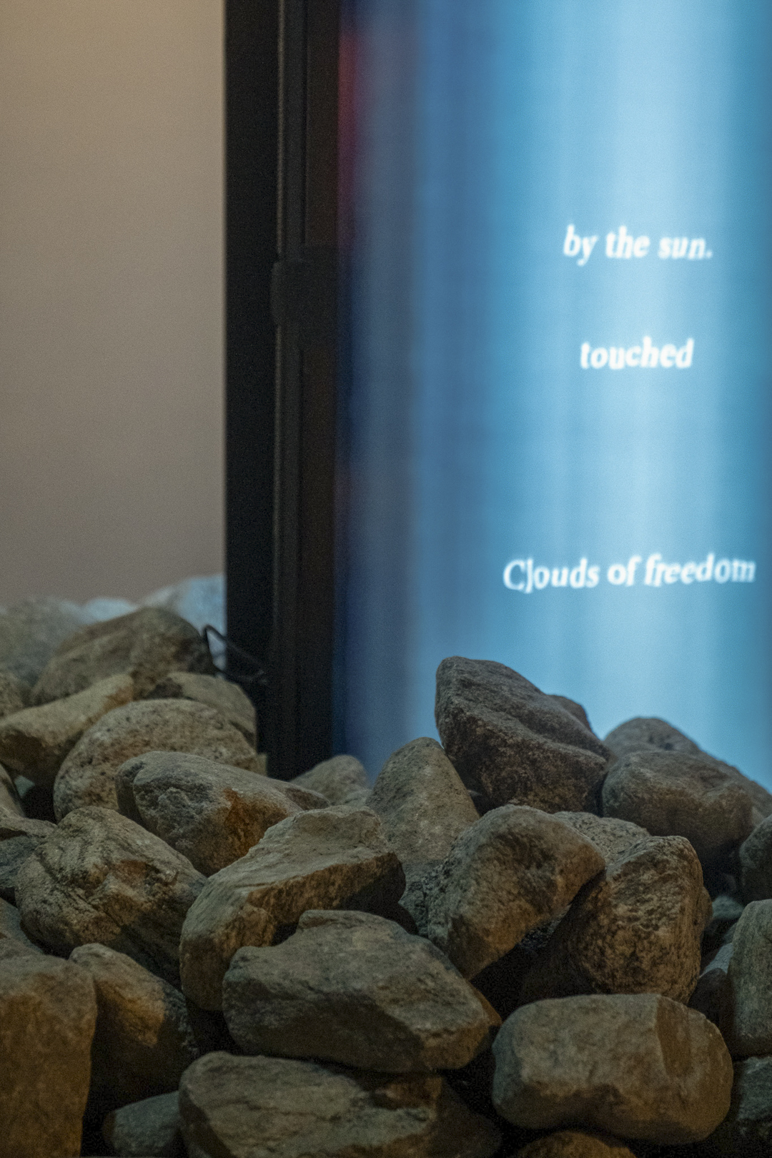 A pile of gray rocks sits in front of a vertical screen displaying blue light and partial white text that reads, by the sun. touched Clouds of freedom.
