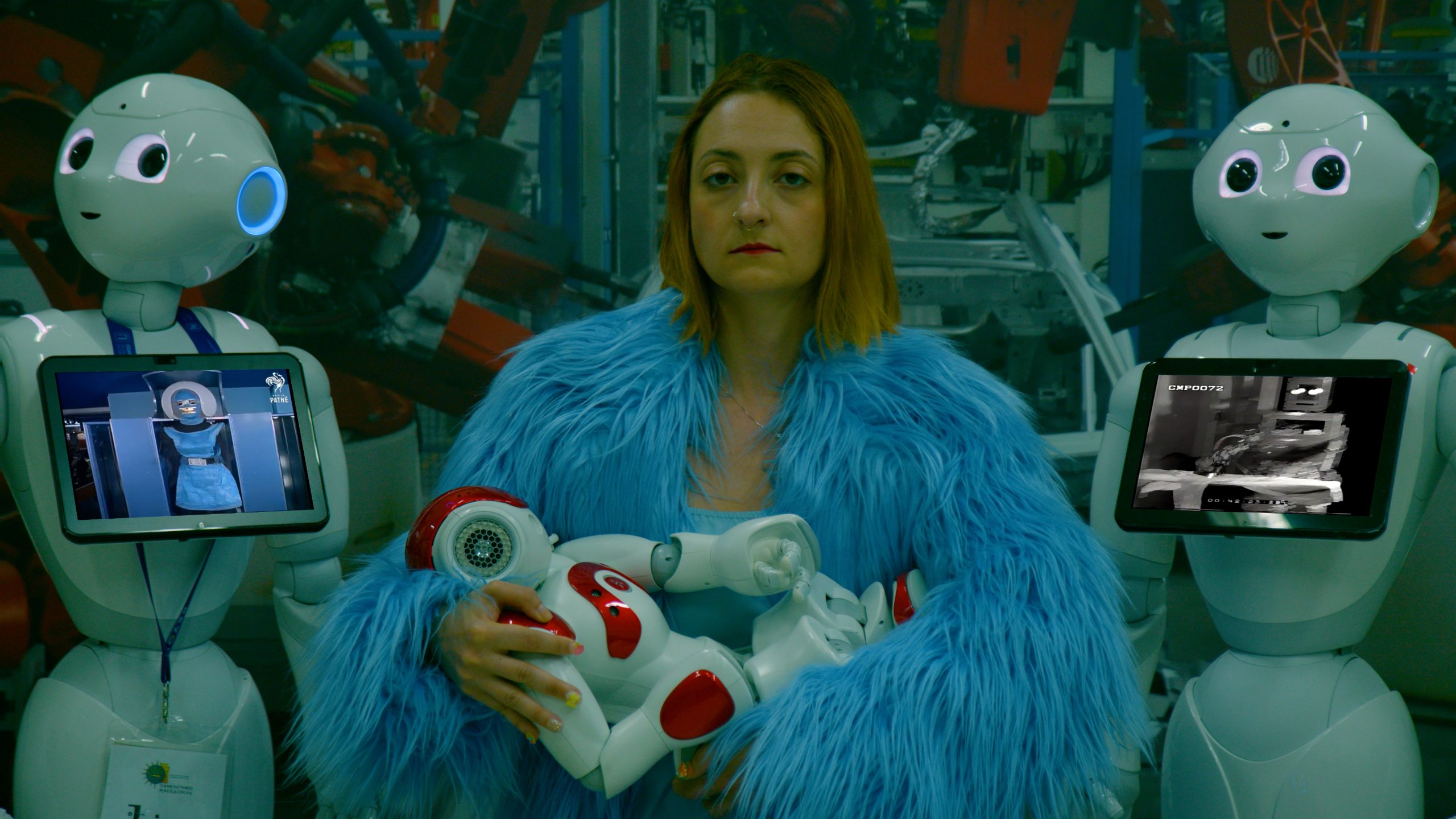 A woman in a blue fur coat holds a red and white robot while standing between two white humanoid robots with screens displaying images in a high-tech, industrial setting.