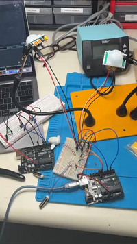 Electronics workbench with Arduino boards, breadboards, jumper wires, sensors, a soldering station, a blue mat, and a laptop in the background, showing an electronics prototyping project in progress.