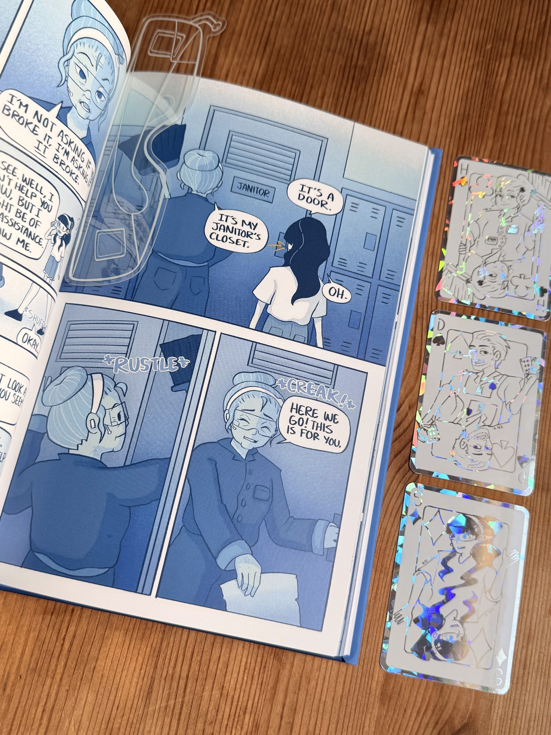 A blue-toned comic book is open on a wooden table next to three collectible holographic cards featuring line art of various characters. A girl and a teacher are depicted in the comic near a Janitor door.