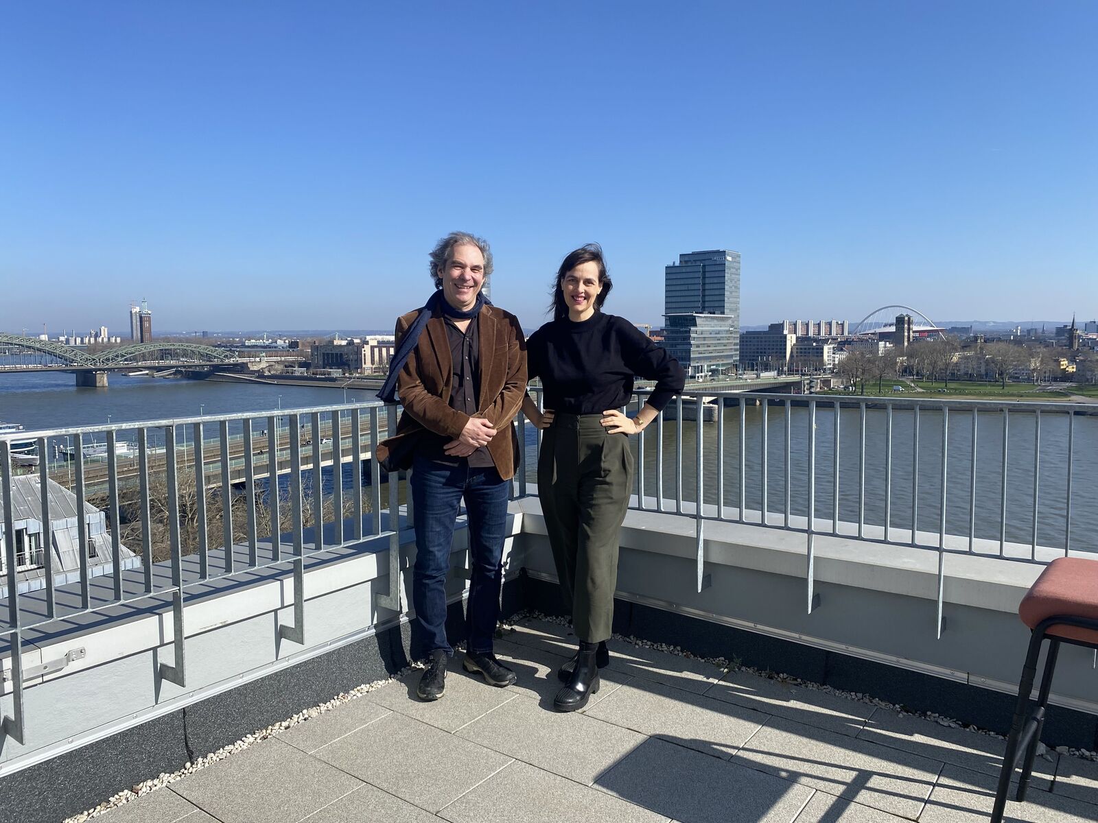 Two people stand on a rooftop terrace with a cityscape, river, and clear blue sky in the background. They are smiling, and metal railings border the terrace.