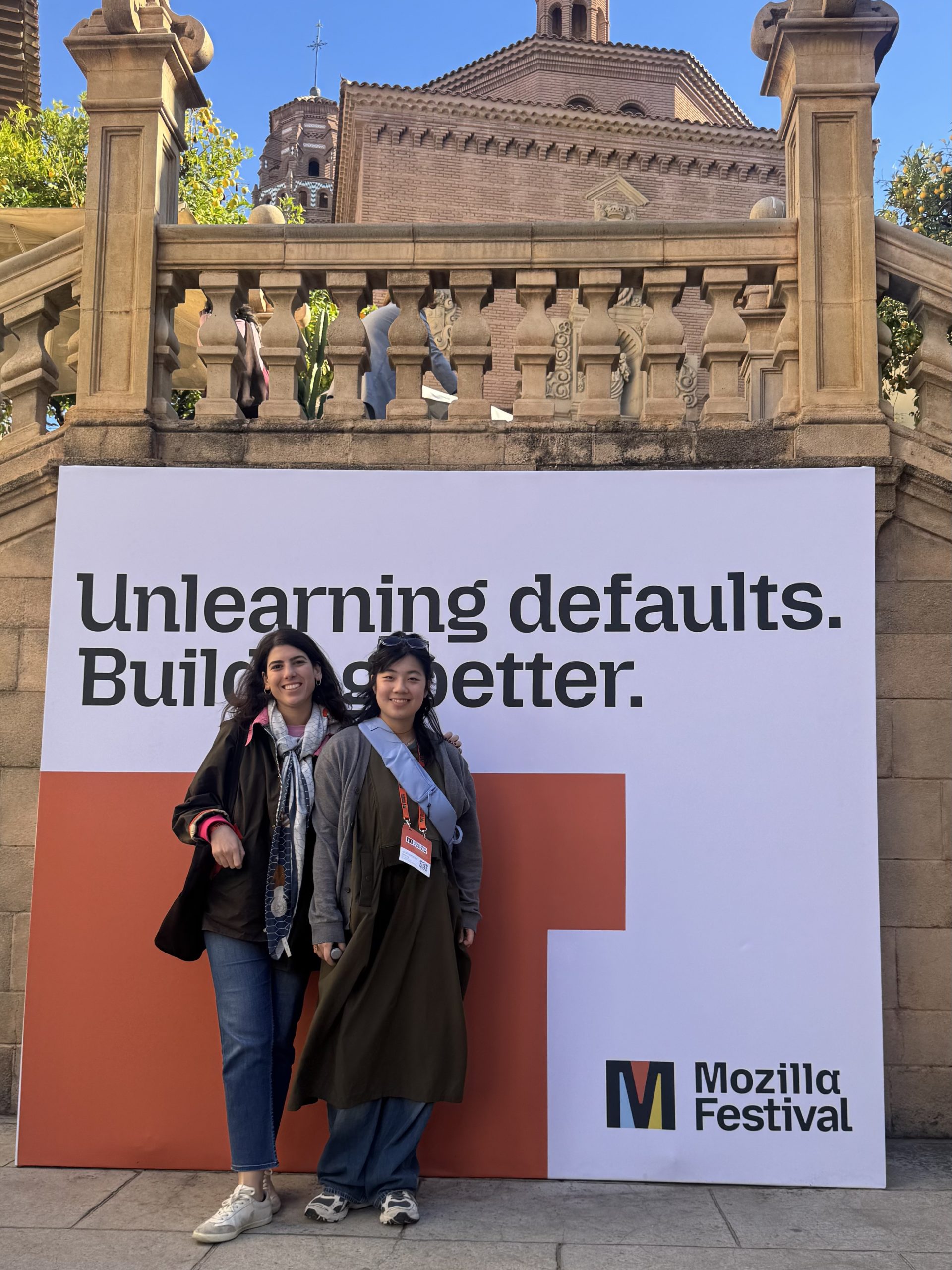 Two people stand and smile in front of a large Mozilla Festival sign that reads “Unlearning defaults. Building better.” Stone steps and a historic brick building appear in the background.