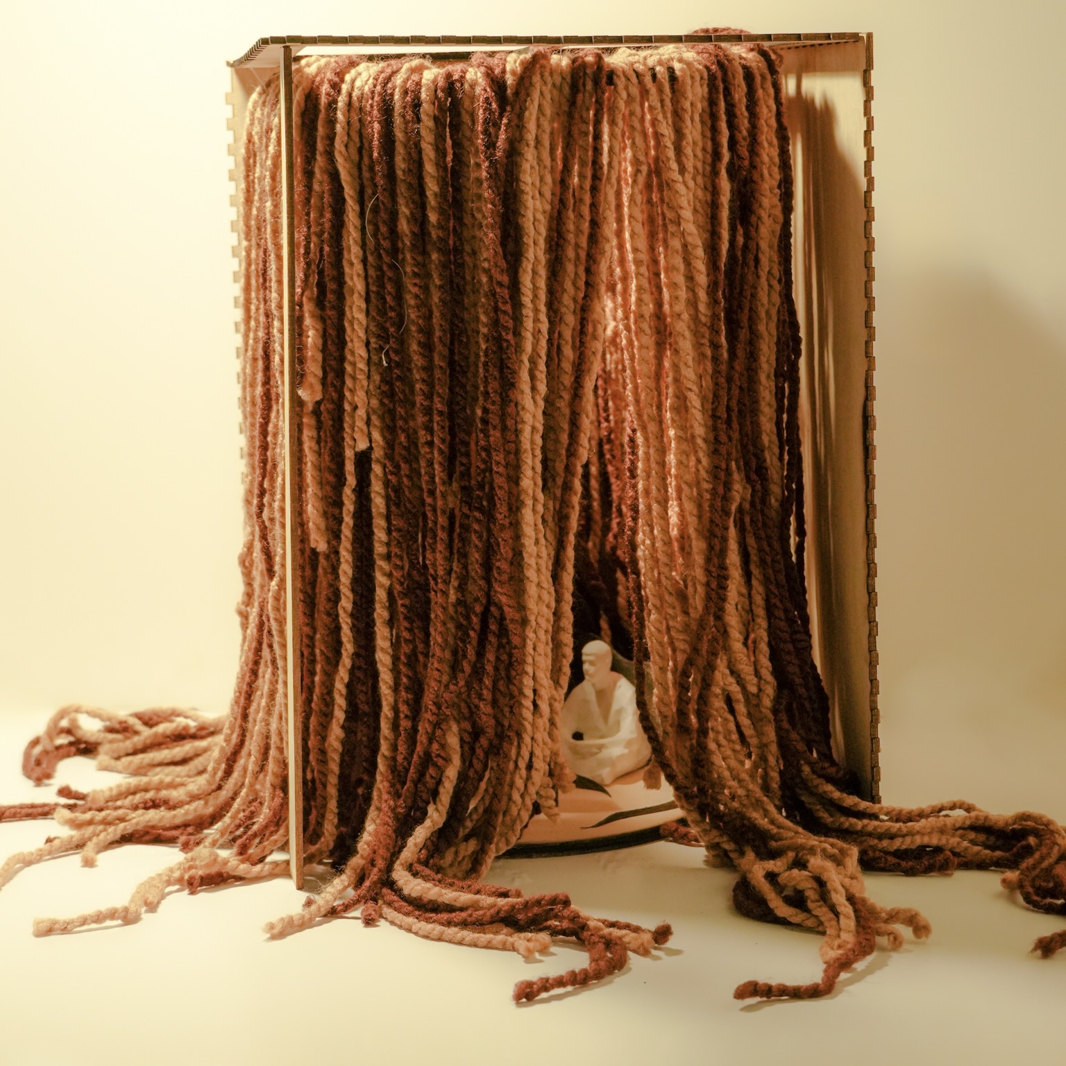 A small white Buddha statue sits inside a wooden box with thick, textured, brown and tan yarn strands draped over the top and sides, cascading down and spreading onto the surface.
