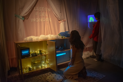 A woman kneels by a glowing cabinet while a person in a red jacket stands by a blue-lit screen. The backdrop reads LISTEN FEEL WONDER, with fabric, clouds, and soft lights creating a dreamy atmosphere.