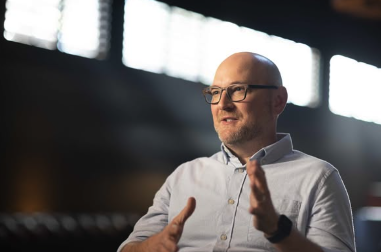 A bald man with glasses and a beard, wearing a light blue button-up shirt, gestures with his hands while speaking in a softly lit room with bright windows in the background.