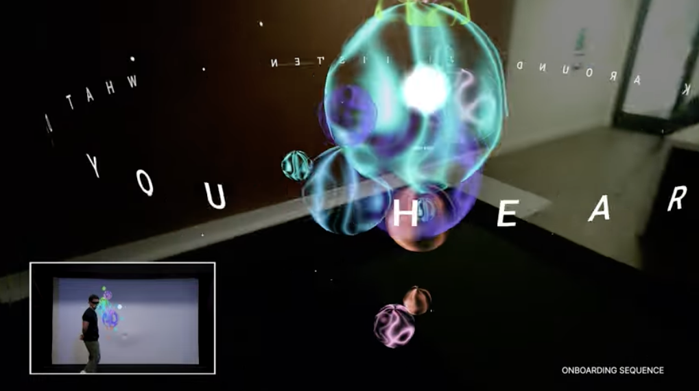 A person stands near a screen displaying floating, colorful, translucent spheres with the words WHAT YOU HEAR among them. A small inset shows the person interacting with the projected visuals.