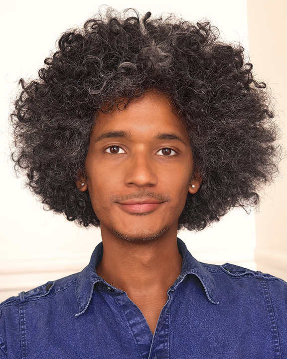 A person with curly, voluminous hair, wearing a denim shirt and small hoop earrings, looks at the camera with a slight smile. The background is softly lit and neutral.
