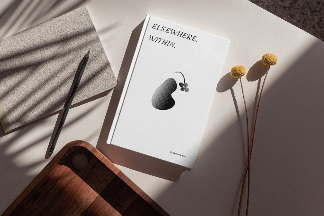 A book titled ELSEWHERE, WITHIN. with a pear illustration on the cover lies on a desk next to a notebook, pencil, wooden tray, and yellow billy ball flowers.