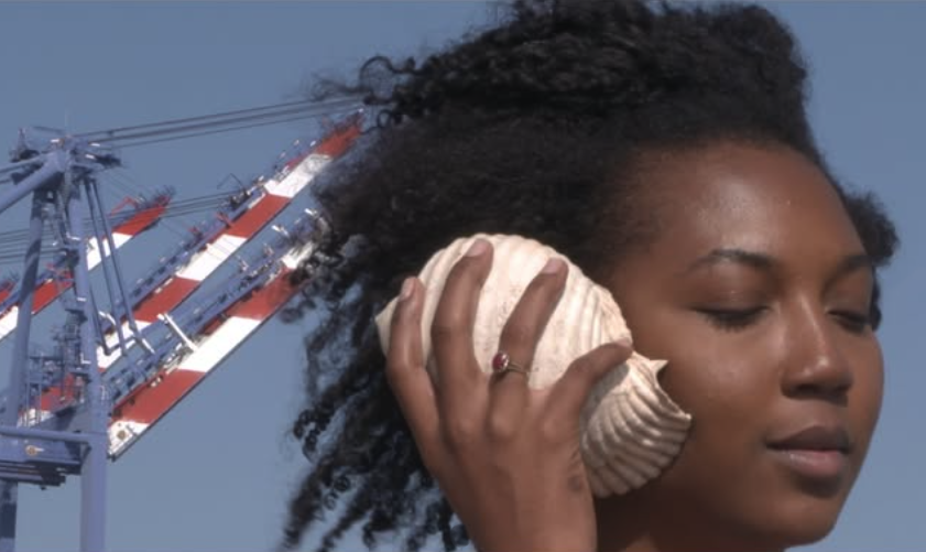 A woman with curly hair holds a large seashell to her ear, eyes closed, standing outdoors with industrial cranes and a blue sky in the background.