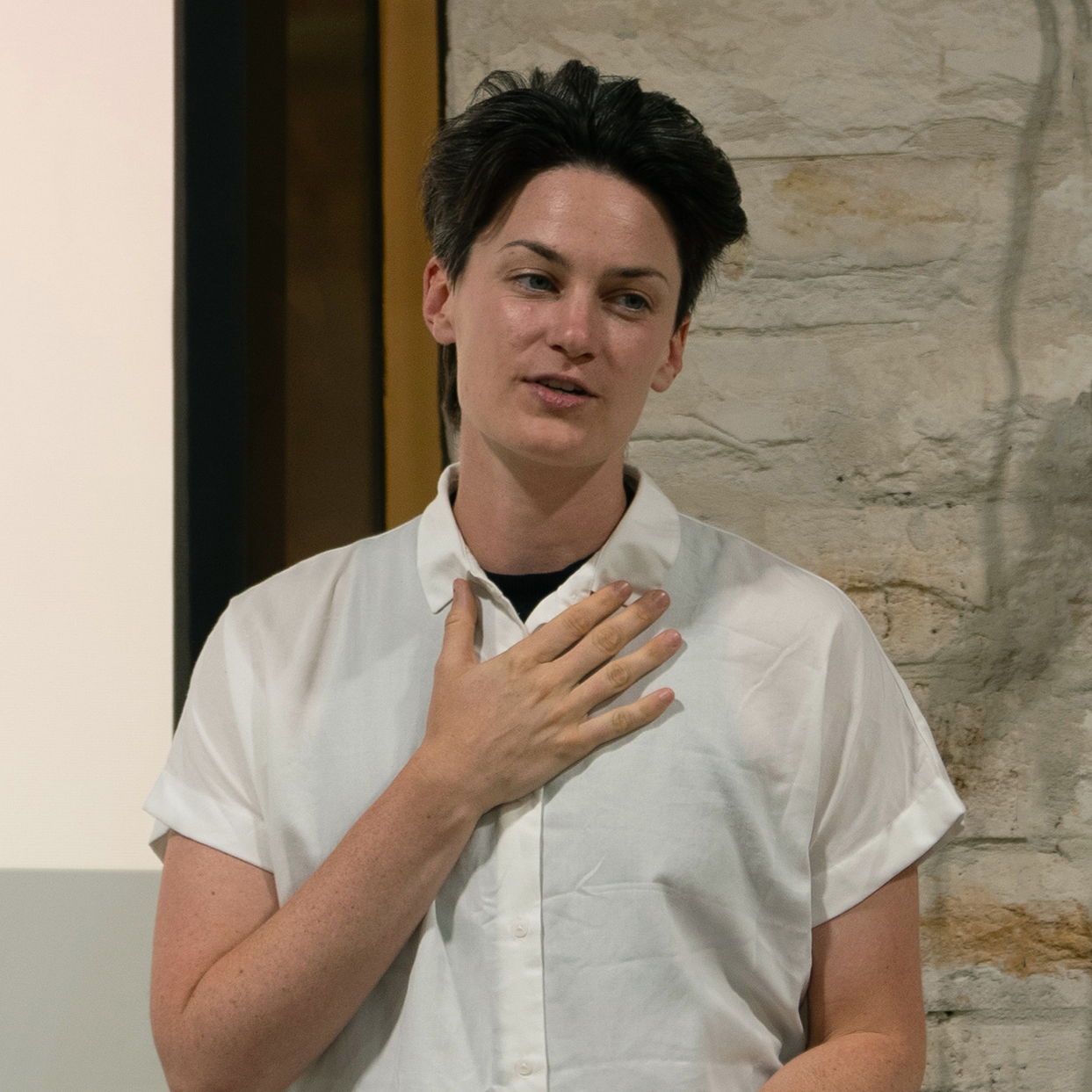 A person with short dark hair wearing a white button-up shirt stands indoors, resting one hand on their chest, appearing to speak or present in front of a stone wall.